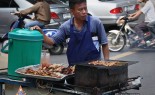 Thai Street Vendor with assorted skewers & sticky rice on a charcoal barbecue cart at a busy intersection.