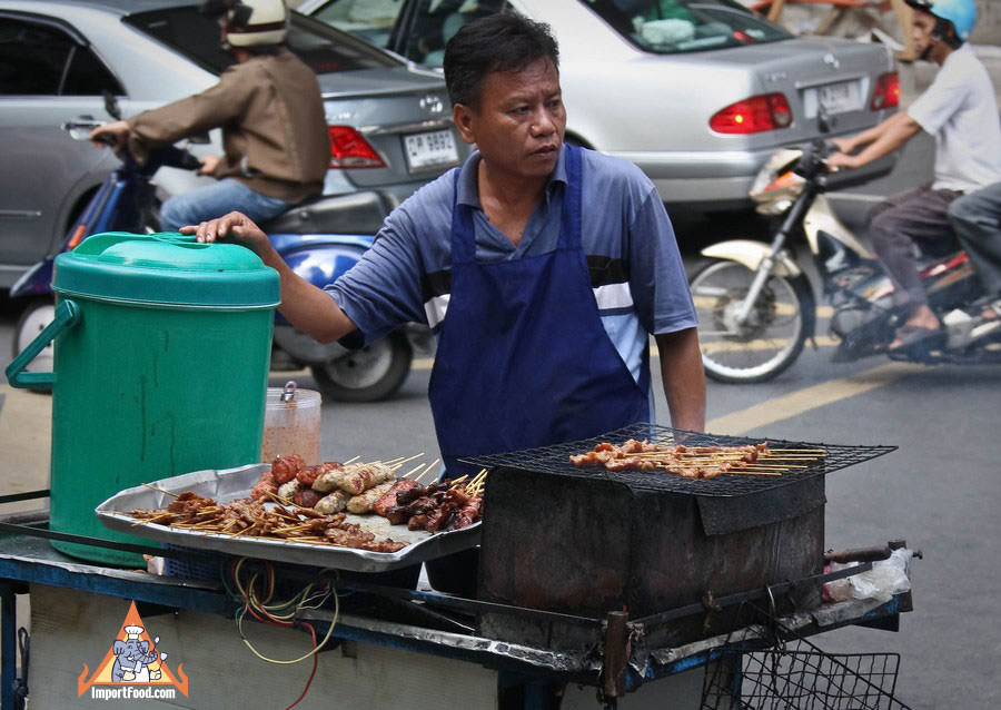 Thai Street Vendor with assorted skewers & sticky rice on a charcoal barbecue cart at a busy intersection.